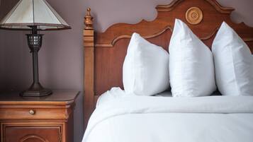 Cama bem arrumada com roupa de cama e almofadas brancas, cabeceira de madeira e mesa de cabeceira com abajur.