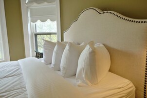 Una cama bien tendida con sábanas y almohadas blancas, un cabecero blanco con adornos de borlas, y una ventana con vistas a árboles.