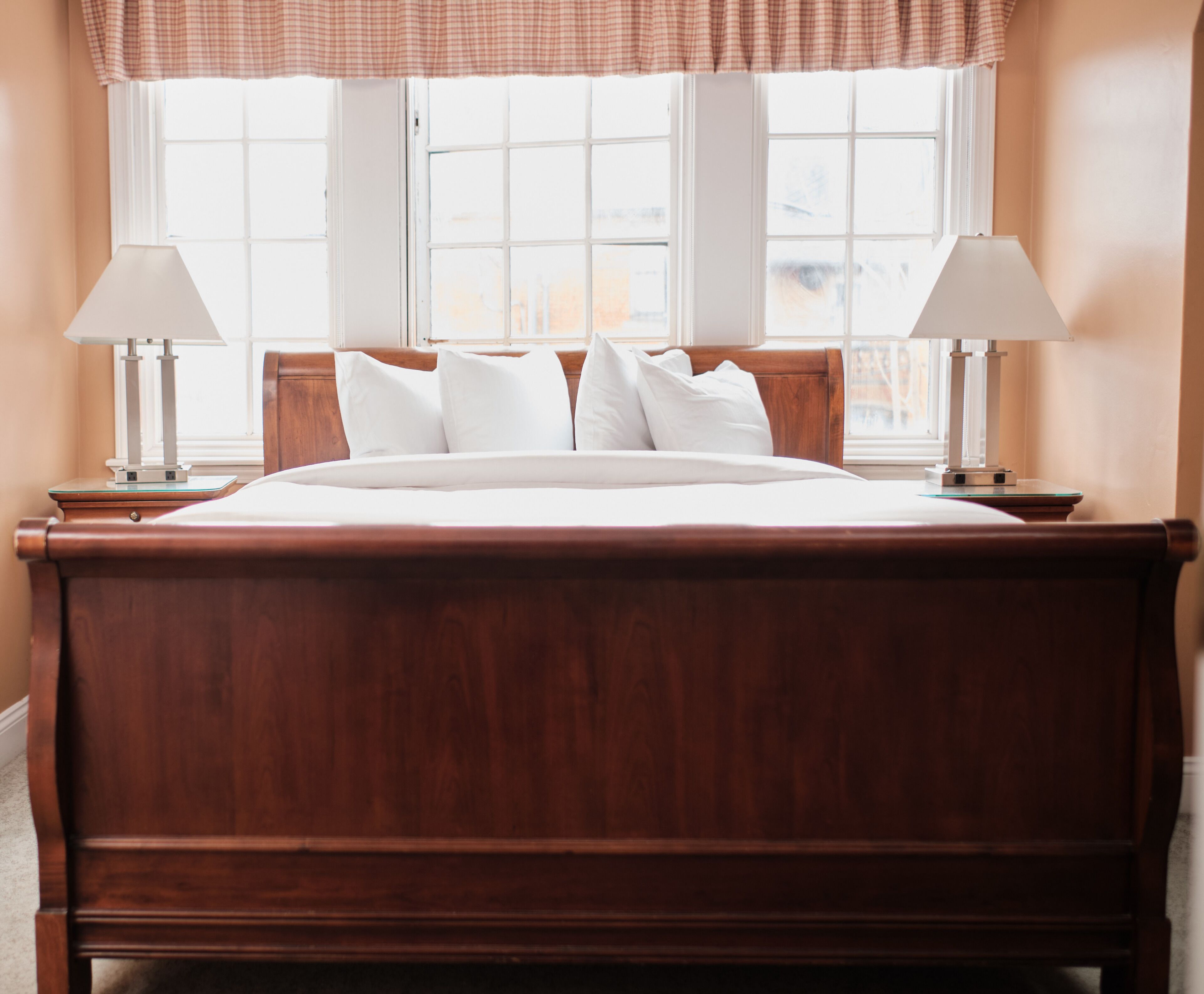 A large wooden bed with white linens, flanked by two bedside tables with lamps, in a room with a window and a plaid valance.