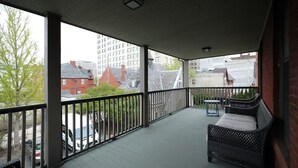 A covered balcony with a wicker sofa and a small table, overlooking a street with buildings and parked cars.