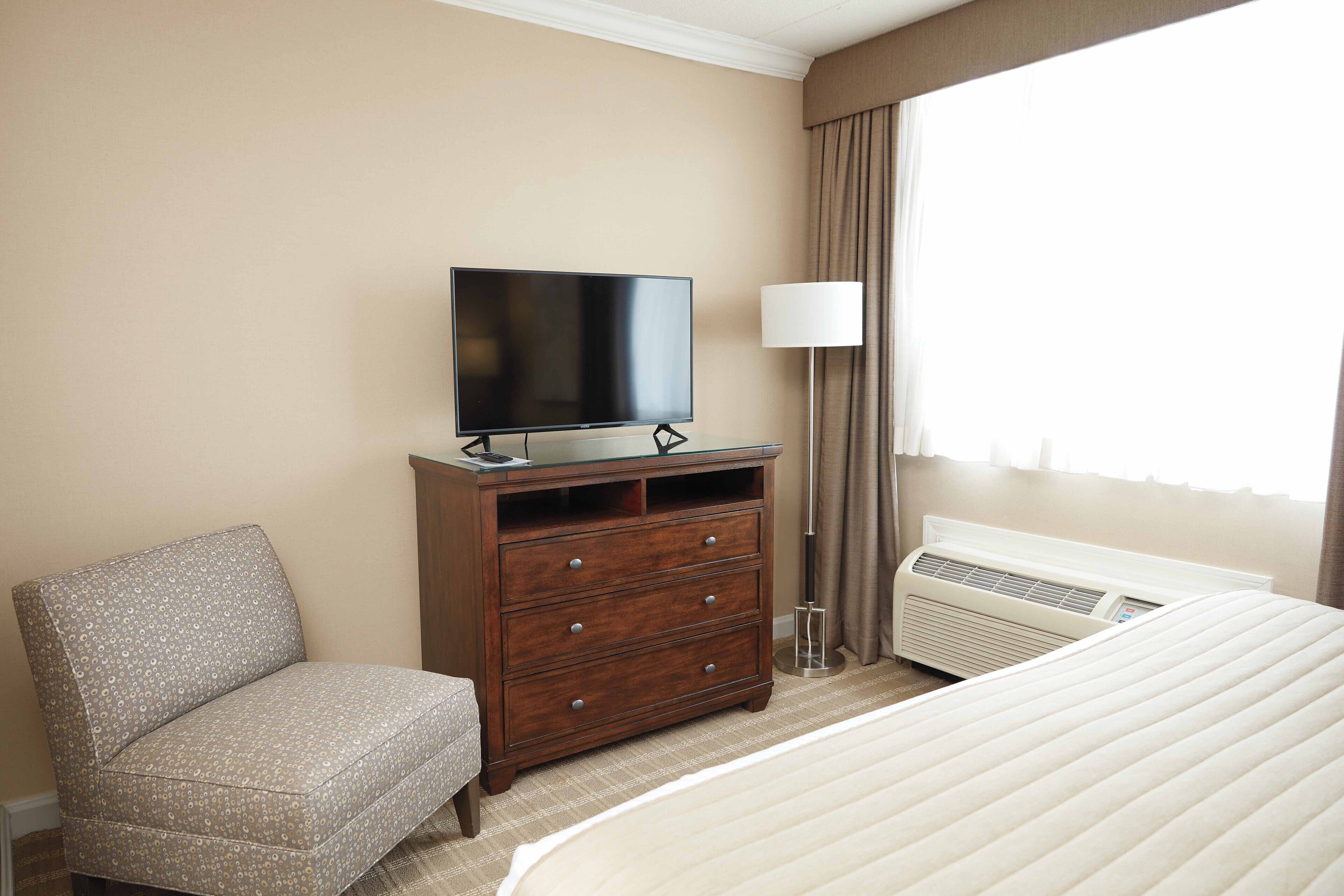 A hotel room with a bed, a TV on a wooden dresser, a chair, a lamp, and an air conditioning unit.