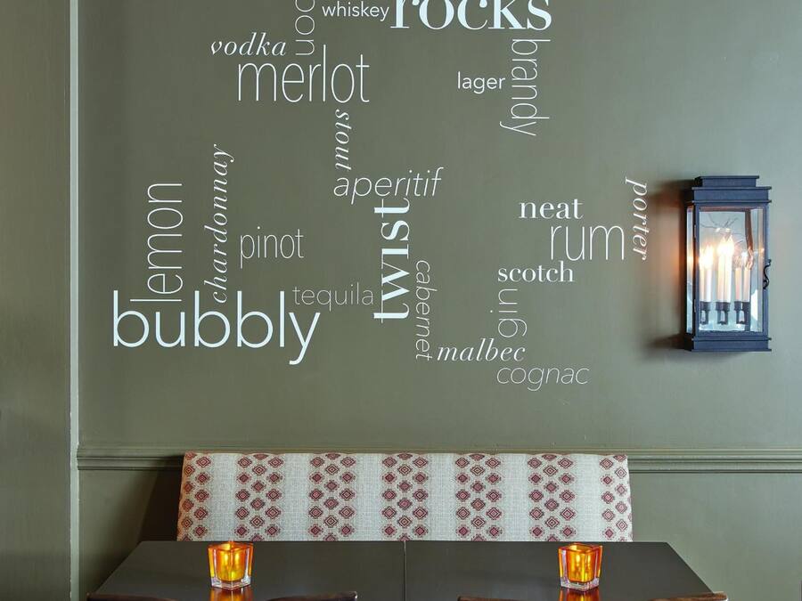 Una pared con una nube de palabras que muestra diferentes tipos de alcohol, una mesa con mantel estampado y una vela encendida sobre la mesa.