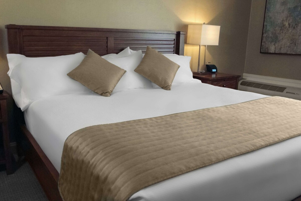 Una cama bien hecha con cabecera de madera, dos mesitas de noche con lámparas, y un cuadro en la pared.