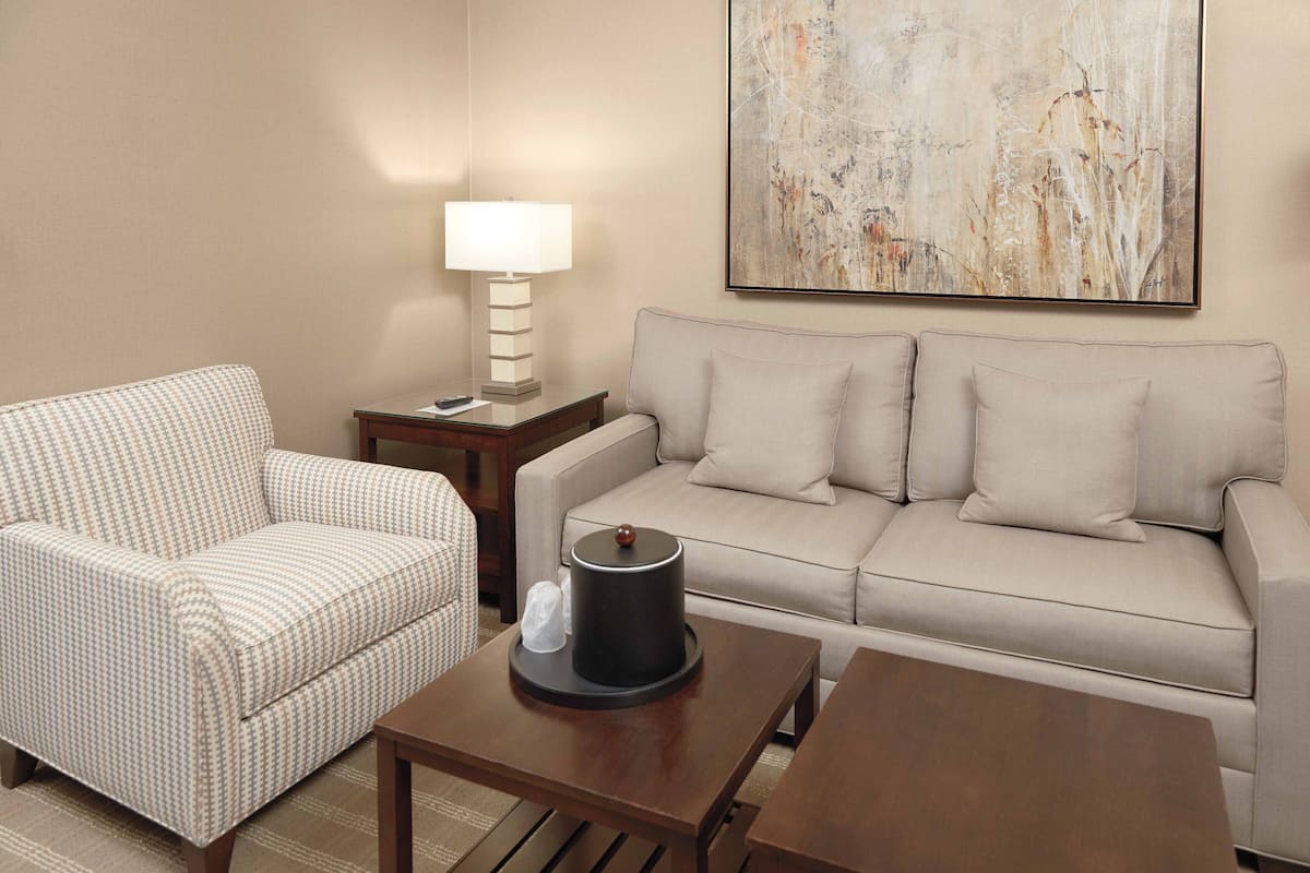 A living room with a checkered armchair, a beige sofa, a coffee table, a side table with a lamp, and a painting on the wall.