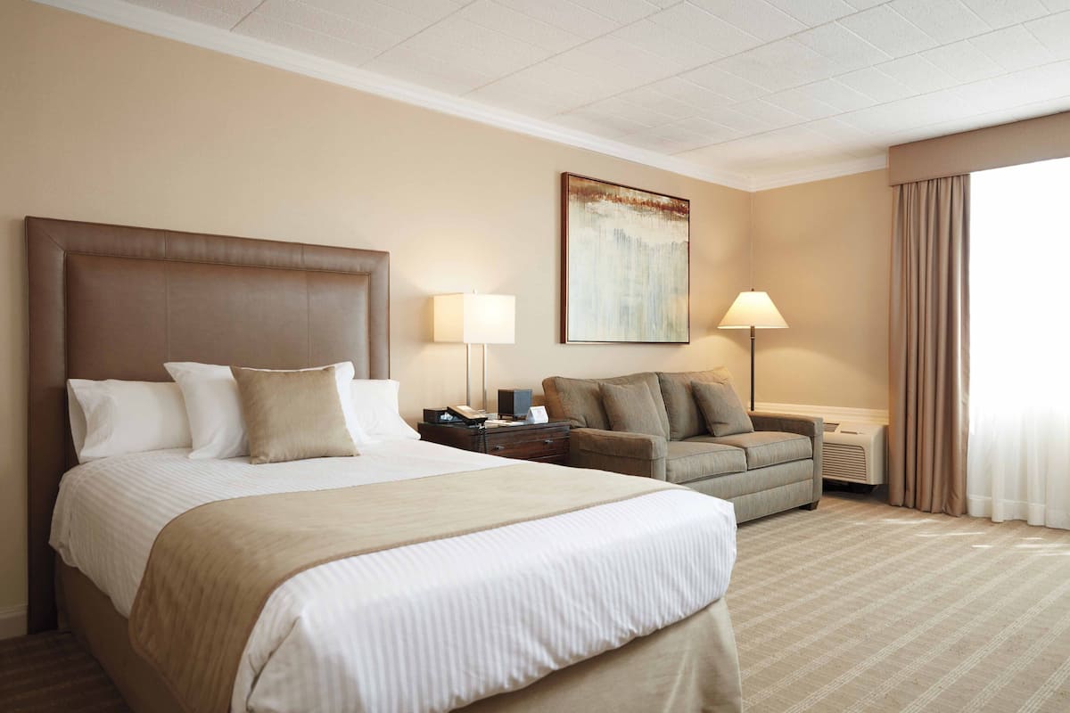 A hotel room with a large bed, a sofa, two bedside lamps, a painting on the wall, and a window with curtains.