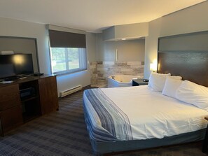 A hotel room with a large bed, a TV, a desk, a bathtub, and a window with a view of greenery.