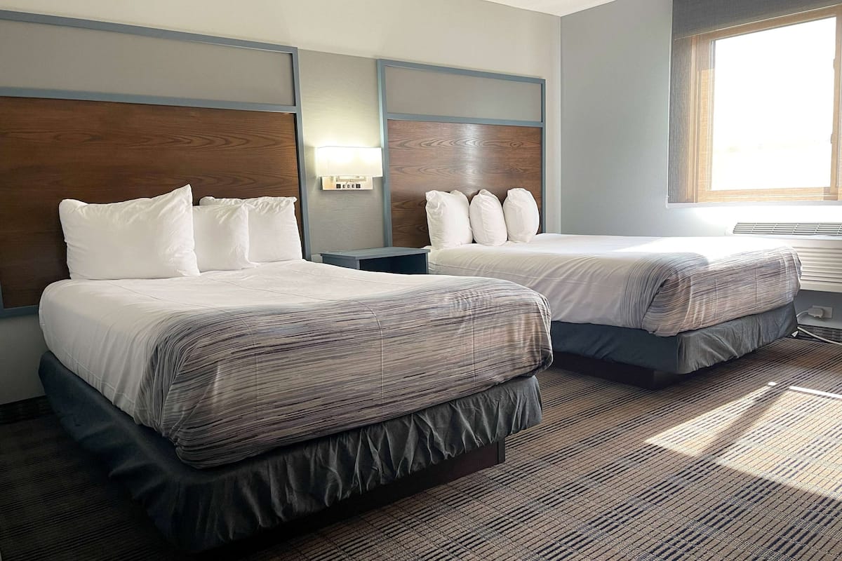 A hotel room with two beds, a nightstand, a lamp, and a window with blinds.
