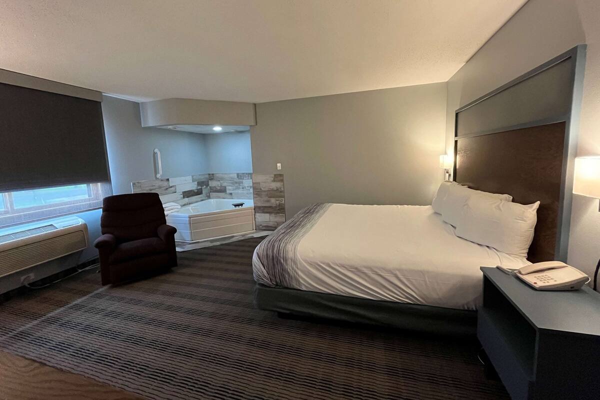 A hotel room with a large bed, a chair, a desk with a phone, a window with a view, and a bathroom with a bathtub.