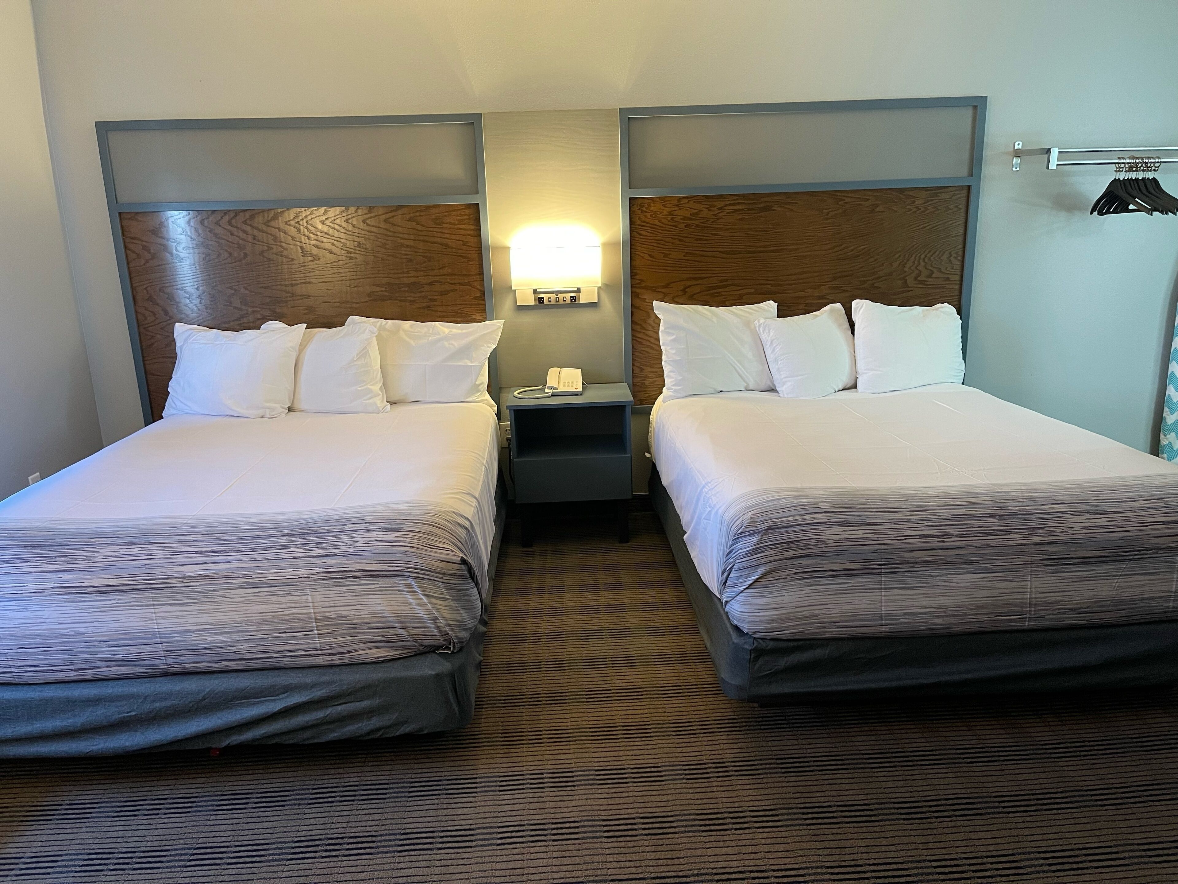 A hotel room with two beds, a nightstand with a phone, and a wall-mounted lamp.