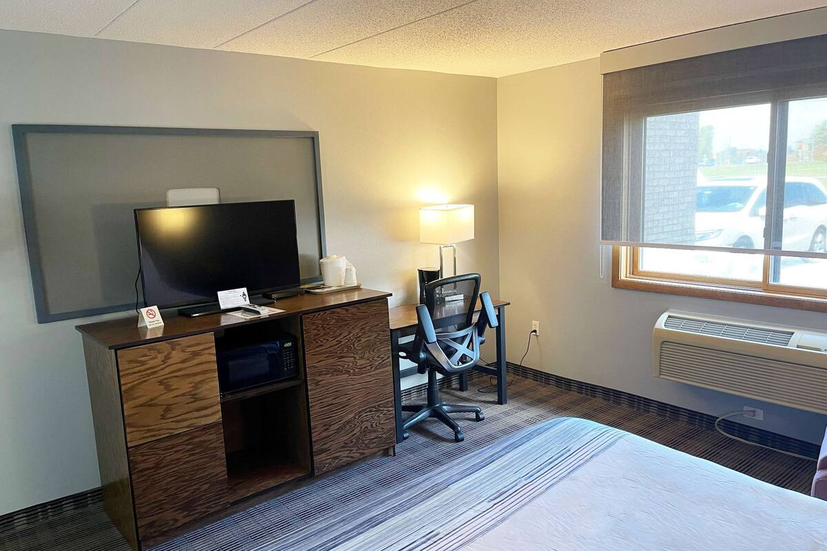 A hotel room with a bed, a desk with a television, a chair, a lamp, and a window with a view of parked cars.