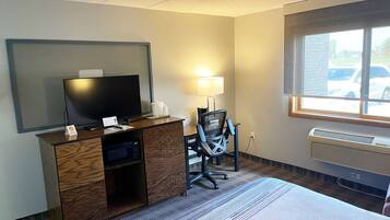 Habitación de hotel con cama, escritorio con televisor, una silla, lámpara y ventana con vista a coches estacionados.