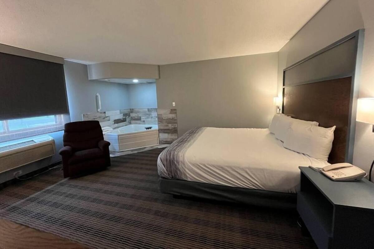 A hotel room with a bed, a chair, a desk with a phone, a window, and a bathroom with a bathtub.