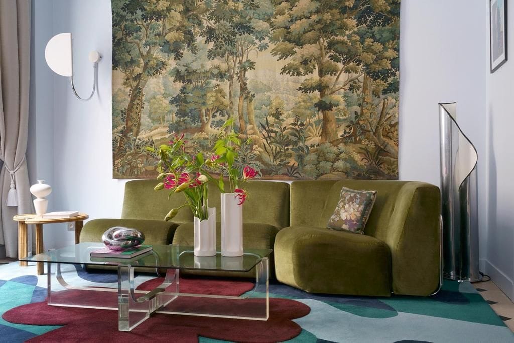 Un salon avec un canapé vert, une table basse en verre et un grand tableau représentant une forêt.