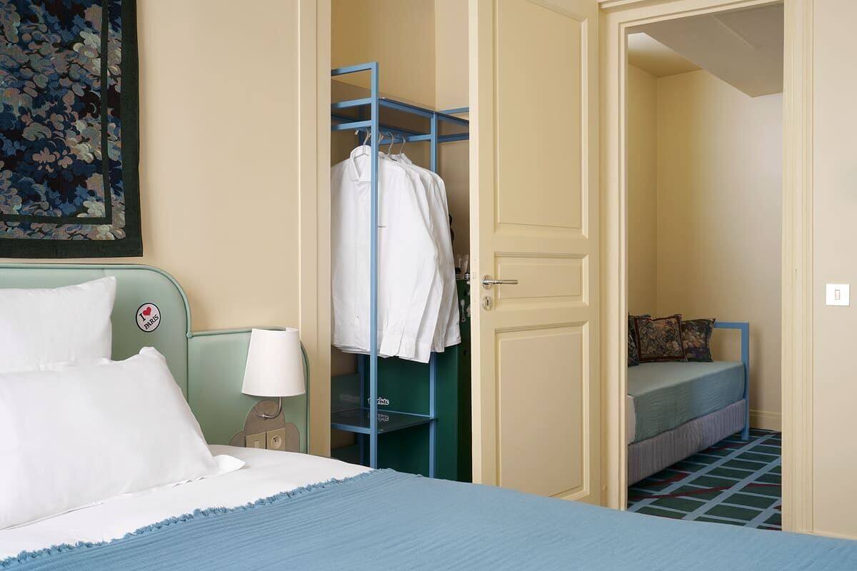 A hotel room with a bed, a wardrobe, a bedside table with a lamp, and a small seating area with a bench and pillows.