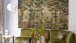 Ein Wohnzimmer mit einem grünen Sofa, einem Glastisch und einem großen Wandteppich mit Waldmotiv.