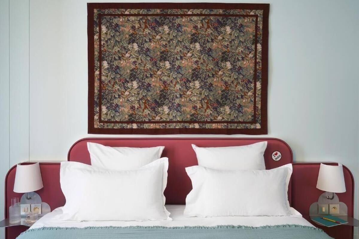 A hotel room with a red headboard, two white pillows, a floral artwork above the bed, and a nightstand with a lamp and a book.