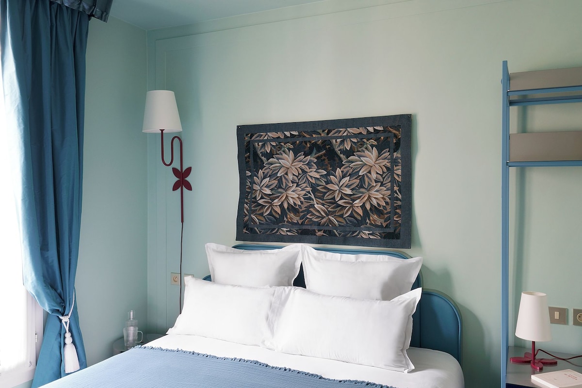 A bedroom with a bed, a nightstand, a lamp, a wall art piece, and a window with curtains.