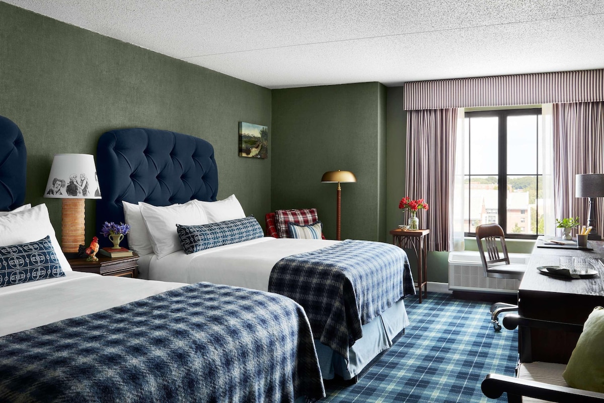 A hotel room with two beds, a desk, a chair, a lamp, and a window with curtains.