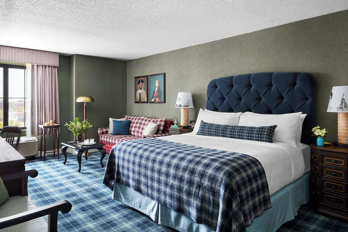 A hotel room with a large bed, a sofa, a small table, and a chair. There is a window with curtains, a lamp, and a picture on the wall.
