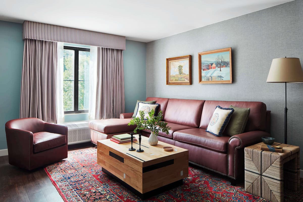 A living room with a leather sofa, a wooden coffee table, a red armchair, a patterned rug, and a large window with curtains.