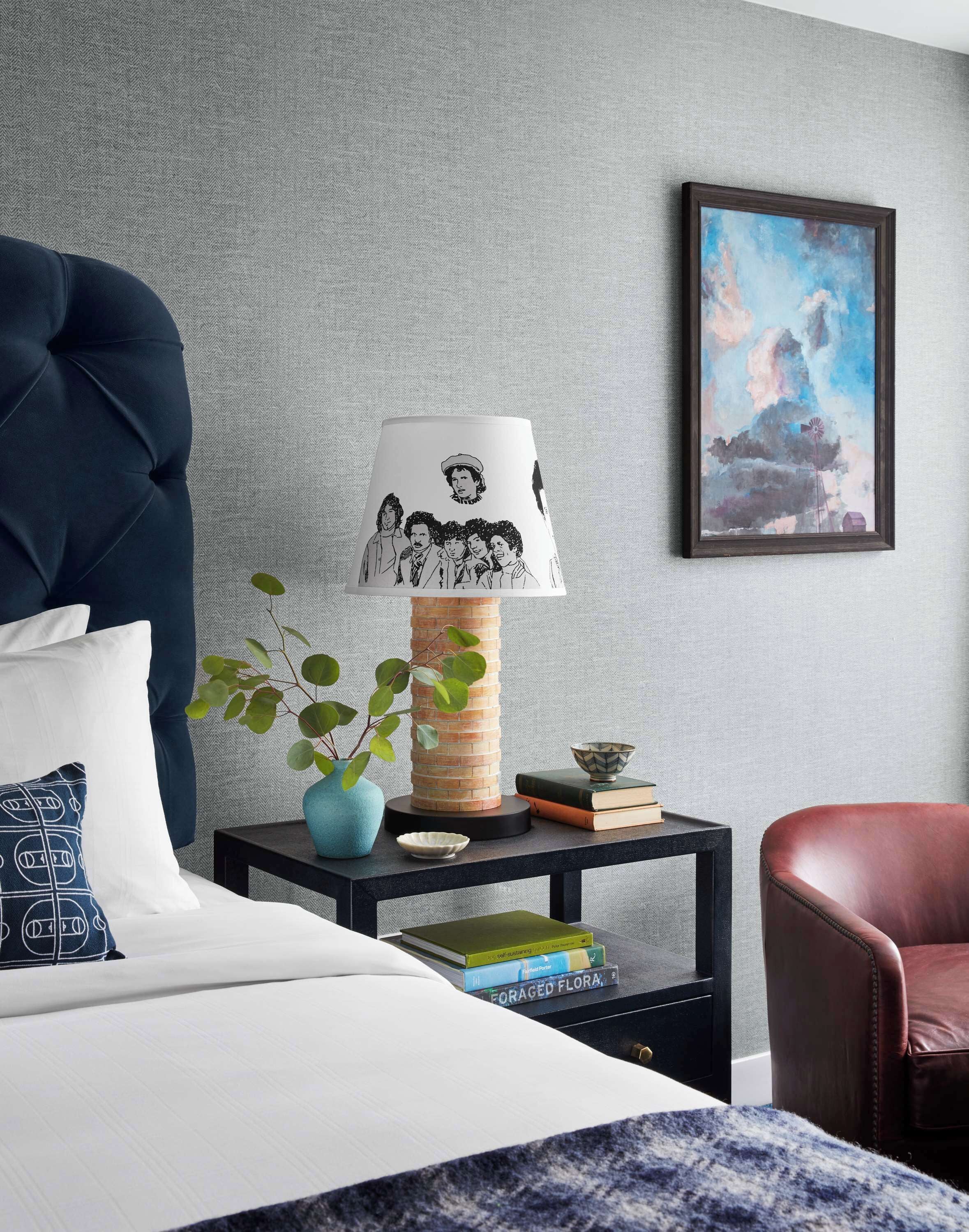 A bedroom with a tufted headboard, a bedside table with a lamp featuring a band illustration, a potted plant, books, and a framed picture on the wall.