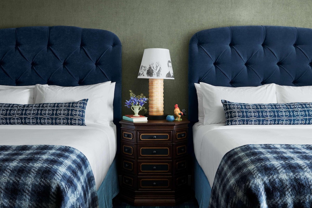 A room with two beds, each with a tufted headboard, a nightstand with drawers, a lamp, and a small figurine.