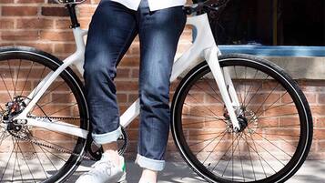 A person standing next to a white bicycle with a black seat and handlebars, wearing jeans and white sneakers.
