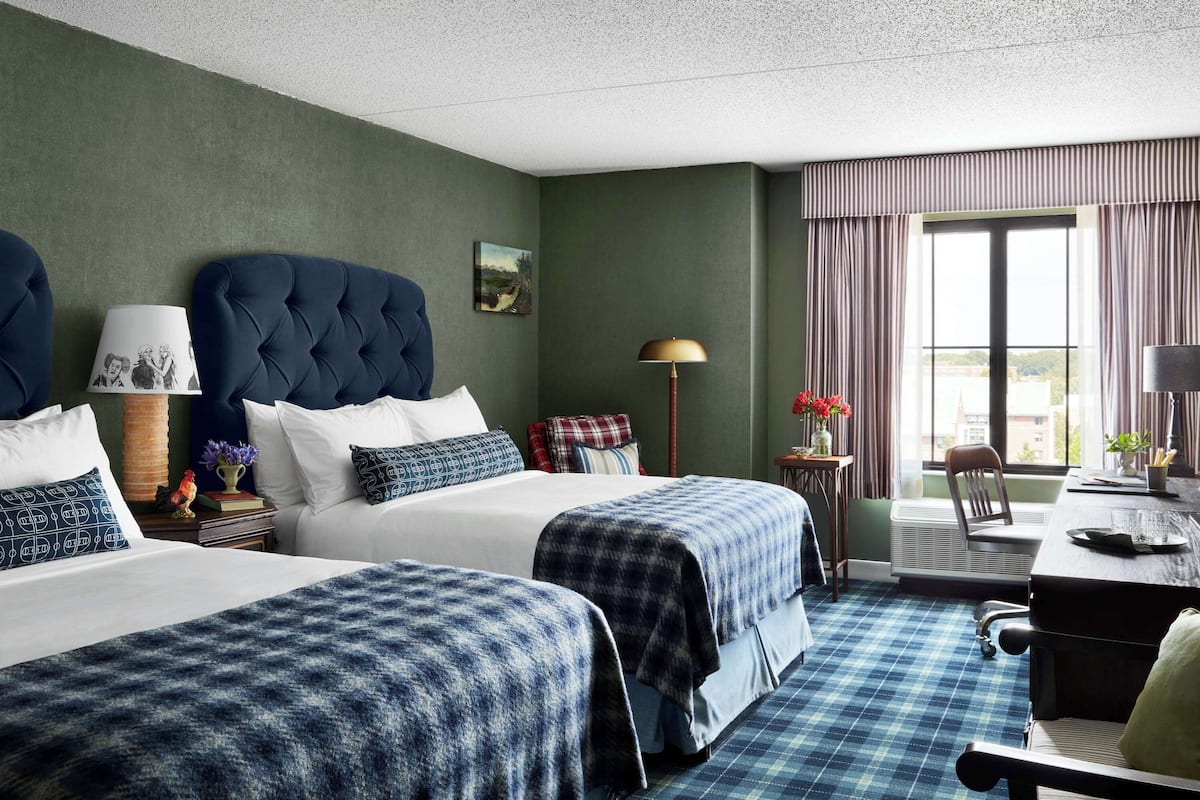 A hotel room with two beds, a desk, a chair, and a window with curtains.