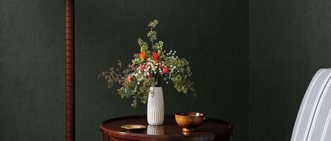 A room with a dark green wall, a round wooden table with a white vase of flowers, a striped armchair, a floor lamp, and a pair of glasses on the armrest.