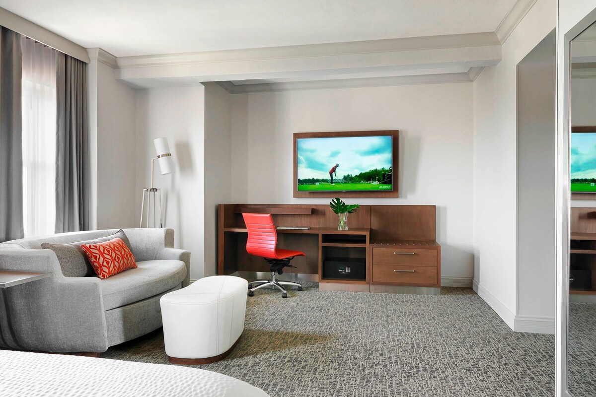 A modern hotel room with a grey sofa, a red office chair, a desk, and a television on the wall.