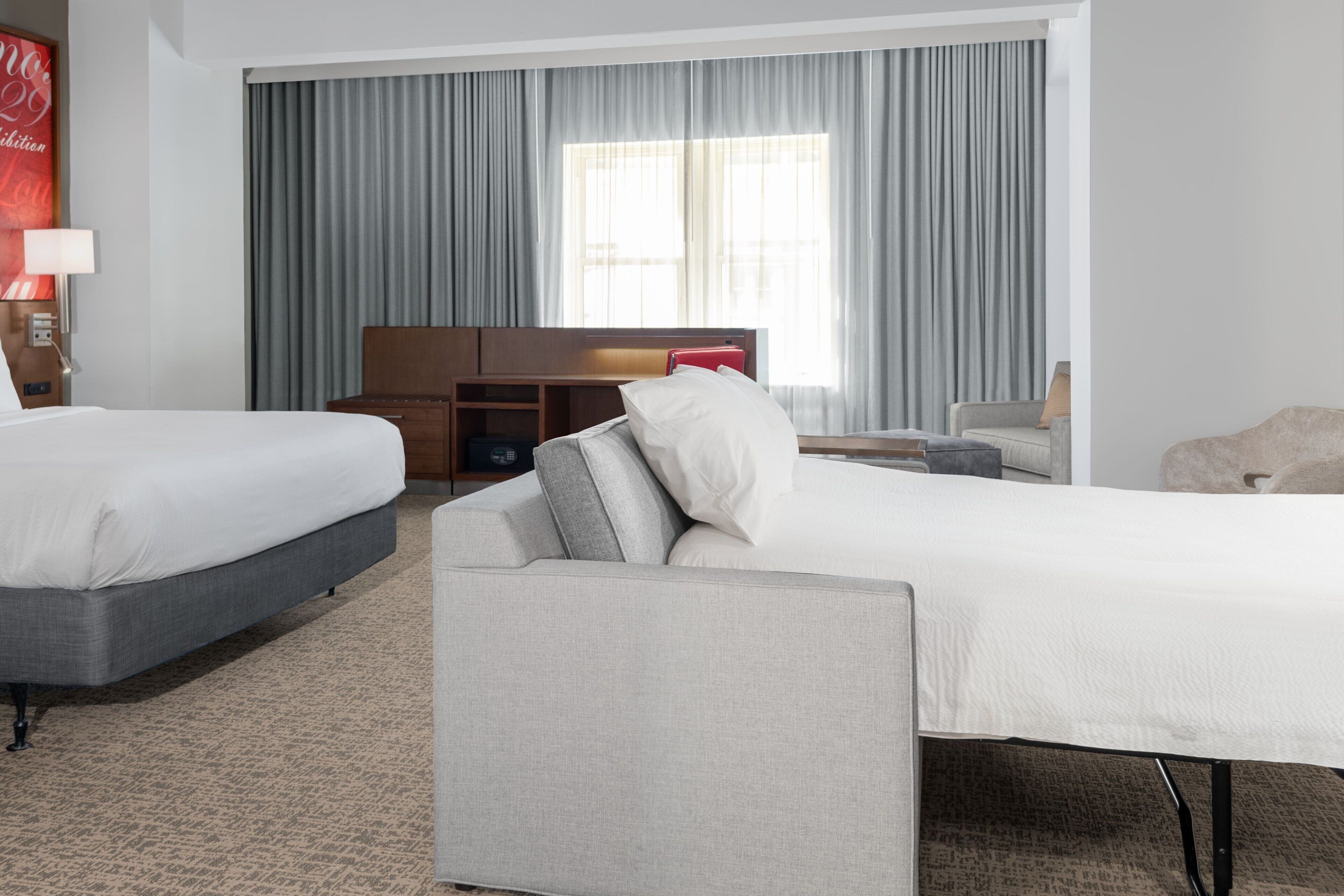 Ein Hotelzimmer mit zwei Betten, einem Sofa, einem Schreibtisch und einem großen Fenster mit Vorhängen.