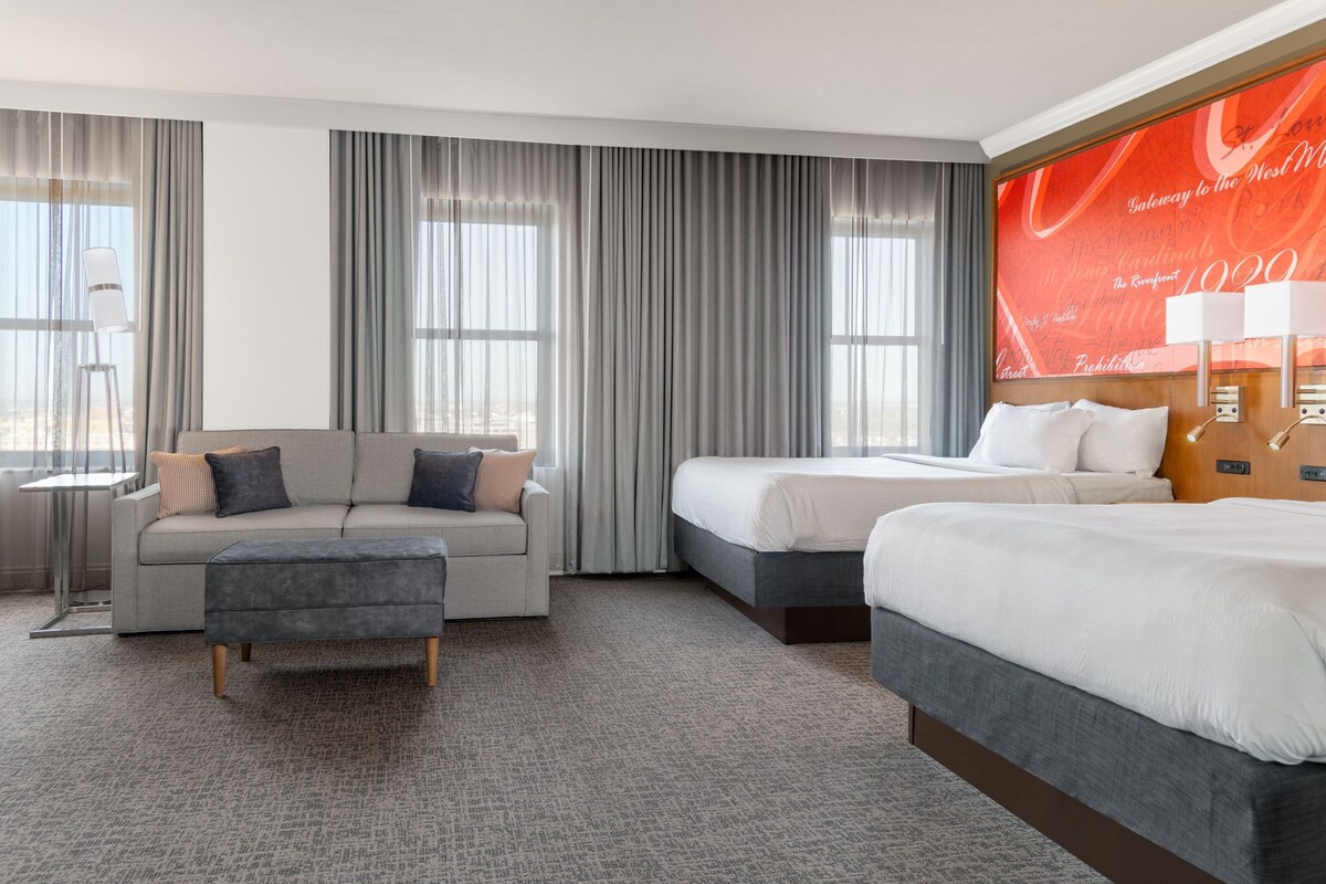 A hotel room with a grey sofa, a grey ottoman, a bed, and a large window with curtains.