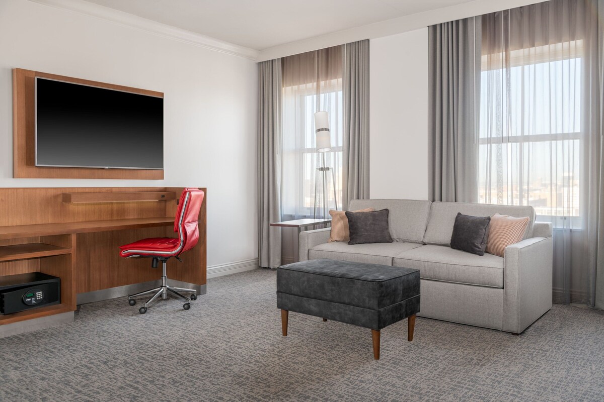 A modern hotel room with a flat-screen TV, a wooden desk, a red office chair, a grey sofa, and a grey ottoman.