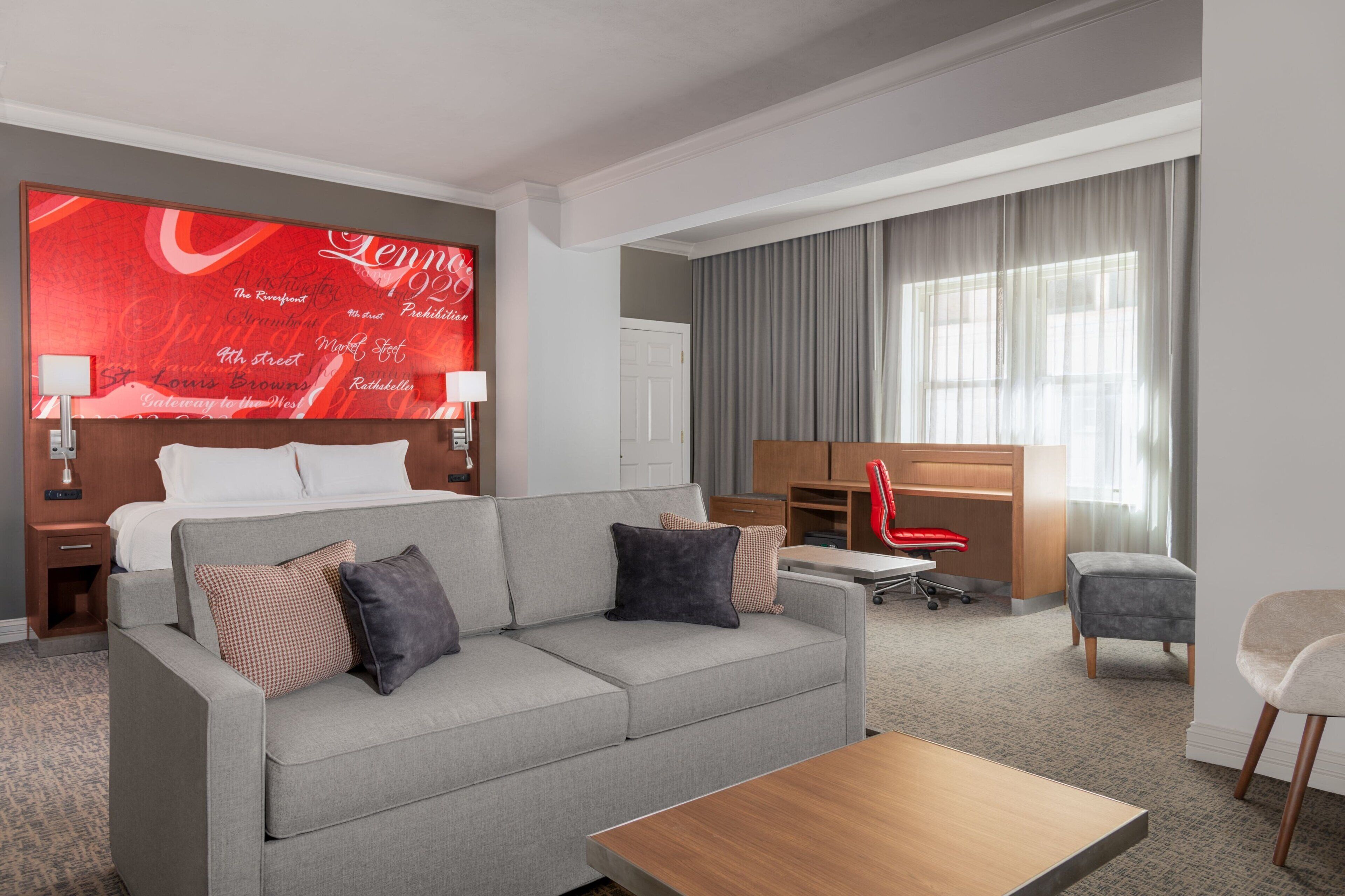 Ein Hotelzimmer mit grauem Sofa, einem Holztisch, einem Schreibtisch mit Stuhl, einem großen Bett und einer roten Wand mit Unterschriften sowie einem Fenster mit Vorhängen.
