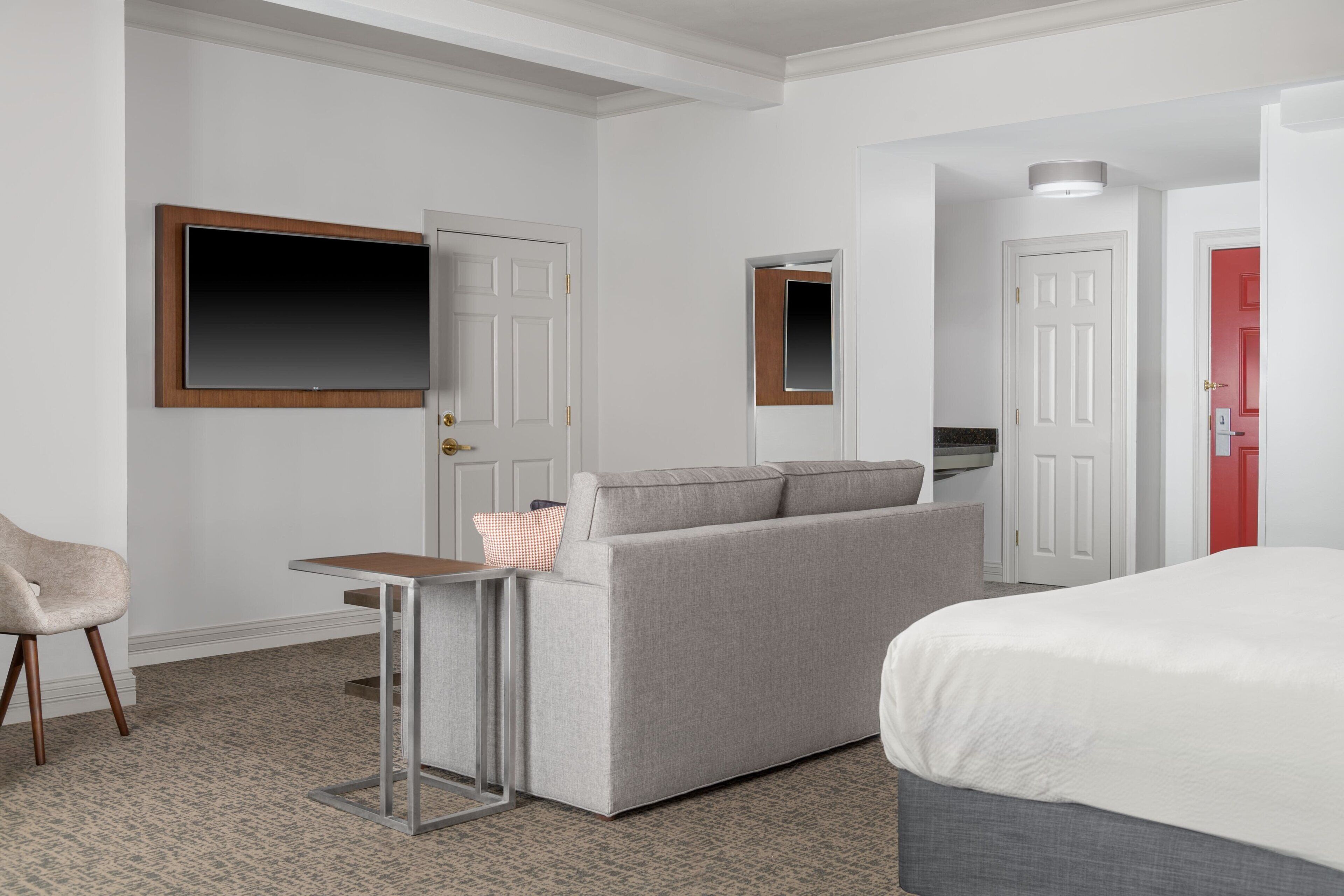 A hotel room with a grey sofa, a small table, a TV mounted on the wall, a bed with a white mattress, and a red door.