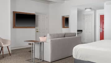 A hotel room with a grey sofa, a small table, a TV mounted on the wall, a bed with a white mattress, and a red door.