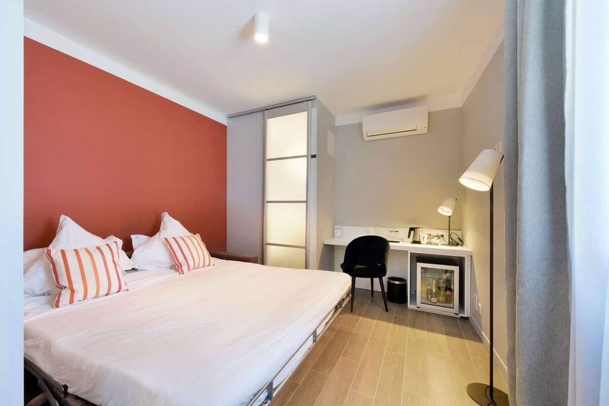 A hotel room with a bed, a desk, a chair, a lamp, and an air conditioning unit.