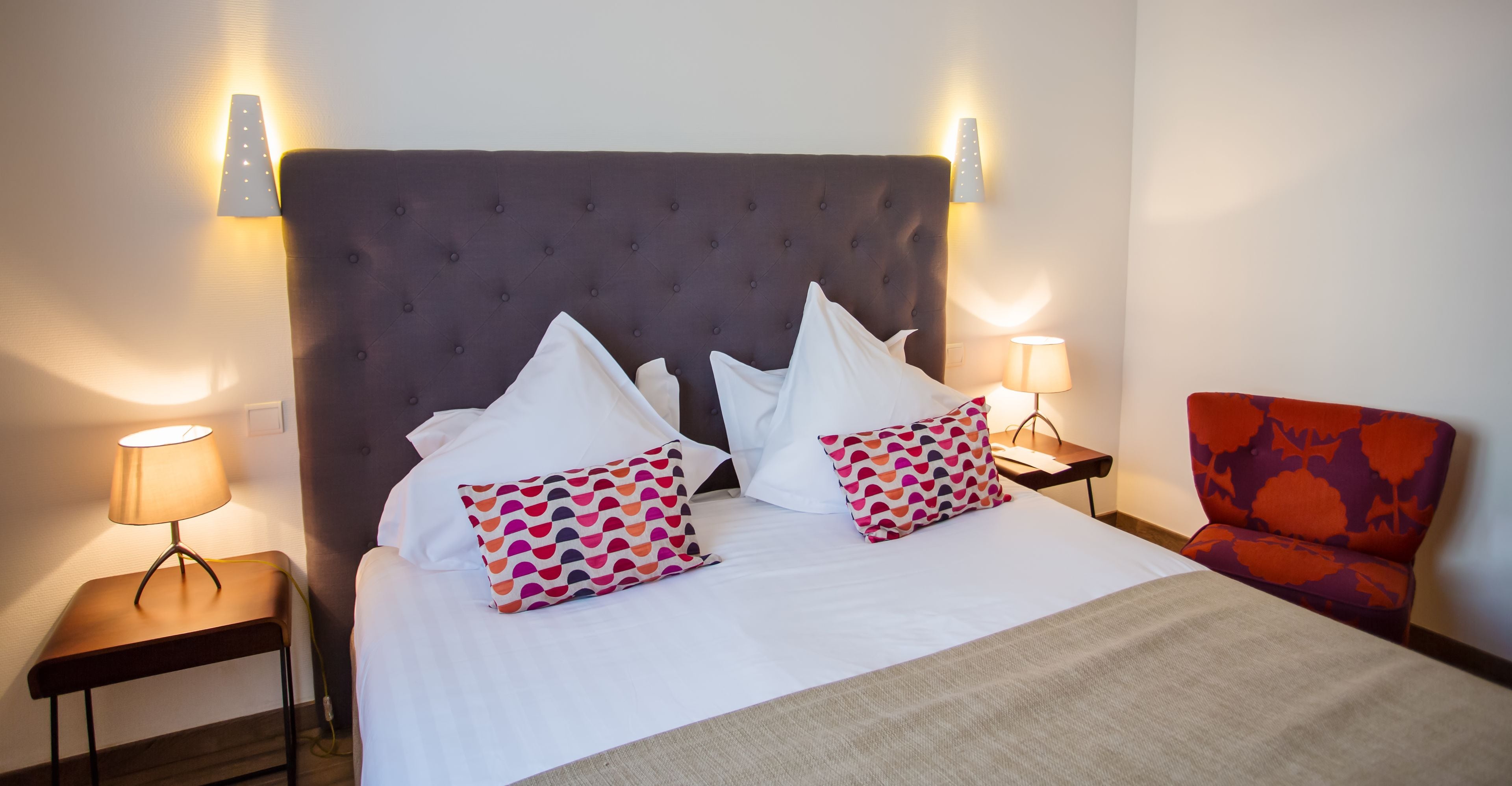 A hotel room with a large bed, two bedside lamps, a chair, and a wall-mounted light fixture.