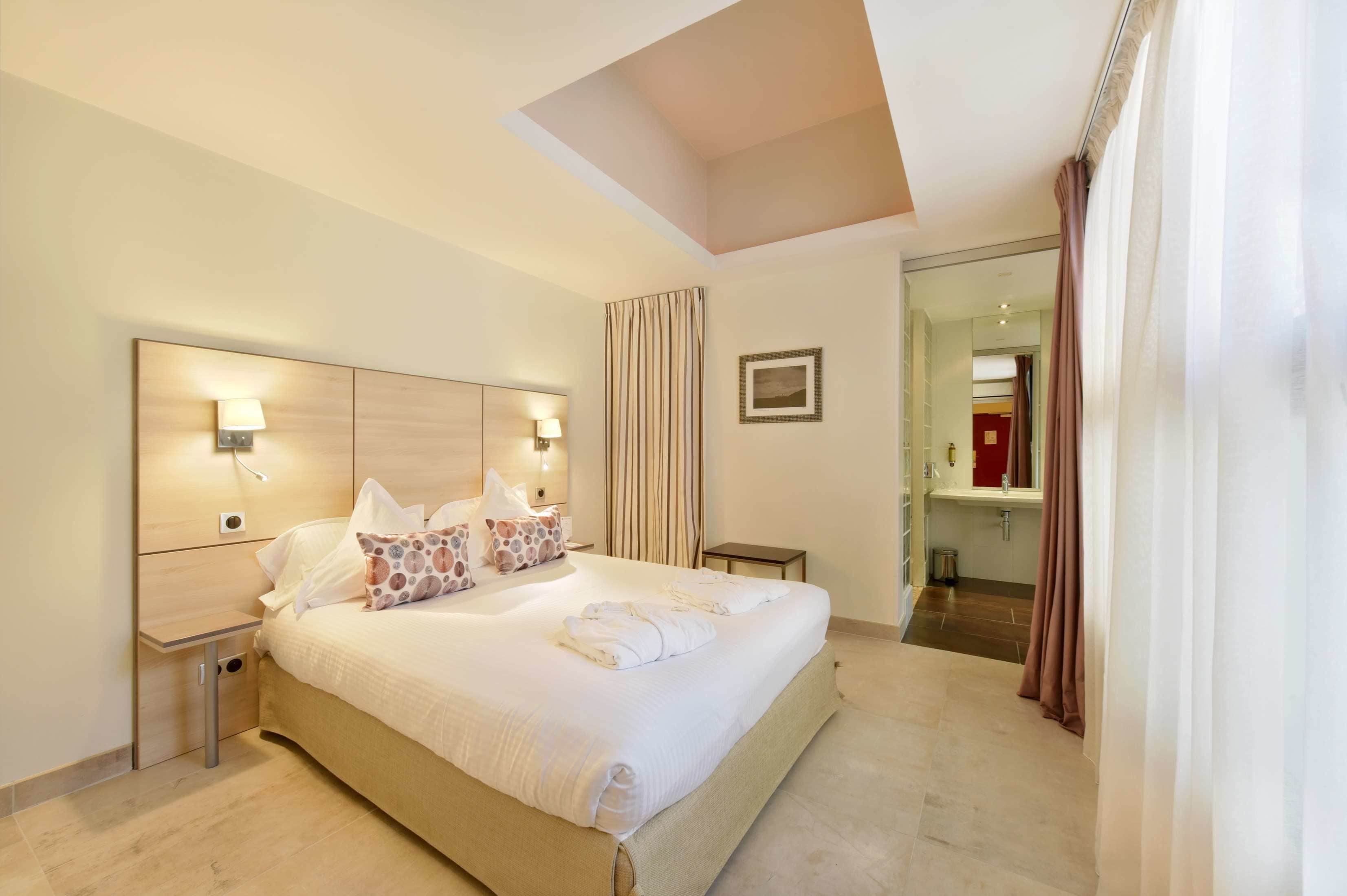 Habitación de hotel moderna con una cama grande, lámparas de noche, una mesita pequeña y vista al baño.
