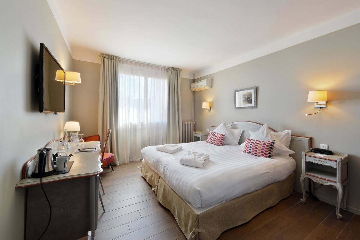 A hotel room with a bed, bedside tables, a desk with a lamp, a TV, and a window with curtains.