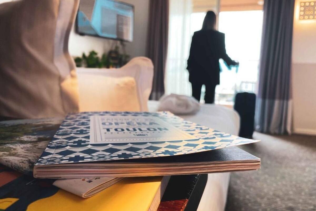A hotel room with a bed, a bedside table with a book, a TV on the wall, and a person walking in the background.