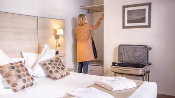 A hotel room with a bed, pillows, a suitcase, a coat rack, and a framed picture on the wall.