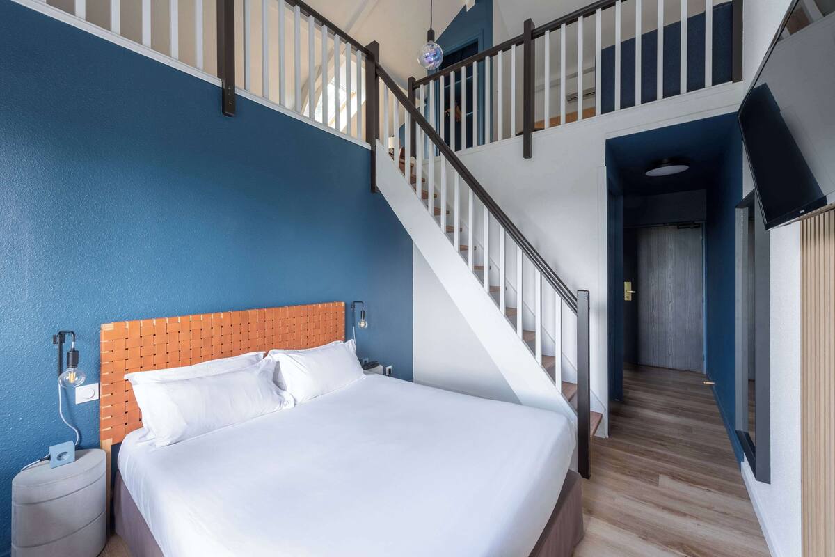 A hotel room with a large bed, a wooden headboard, a nightstand, a wall-mounted lamp, and a staircase leading to an upper level.