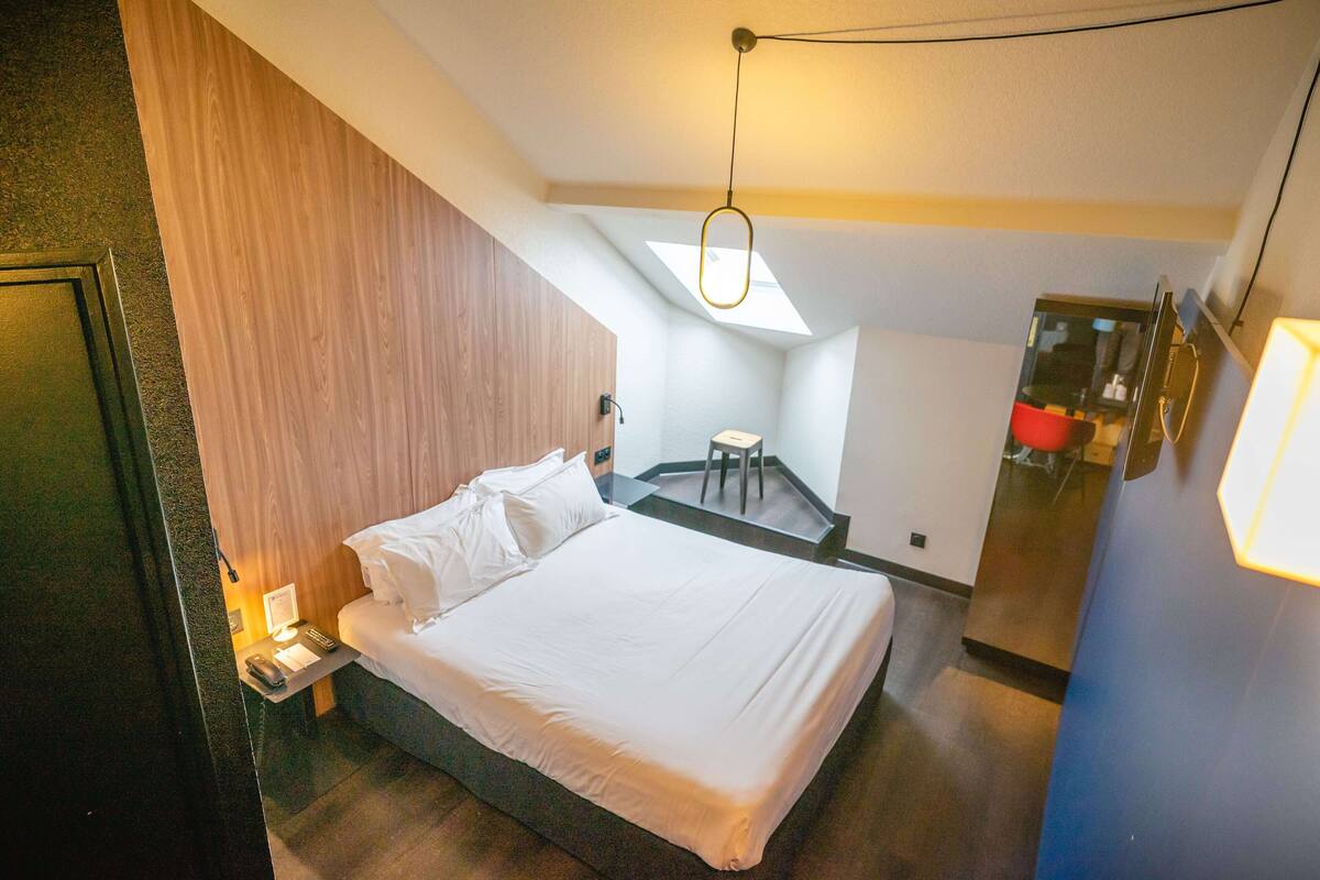 A modern hotel room with a large bed, a desk with a lamp, and a small seating area with a chair.