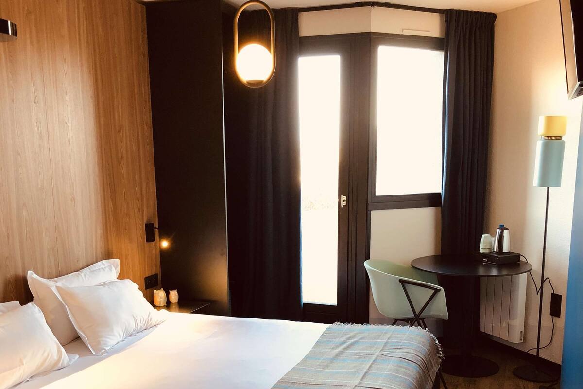 A hotel room with a large bed, a desk, a chair, a lamp, and a view of the outdoors.