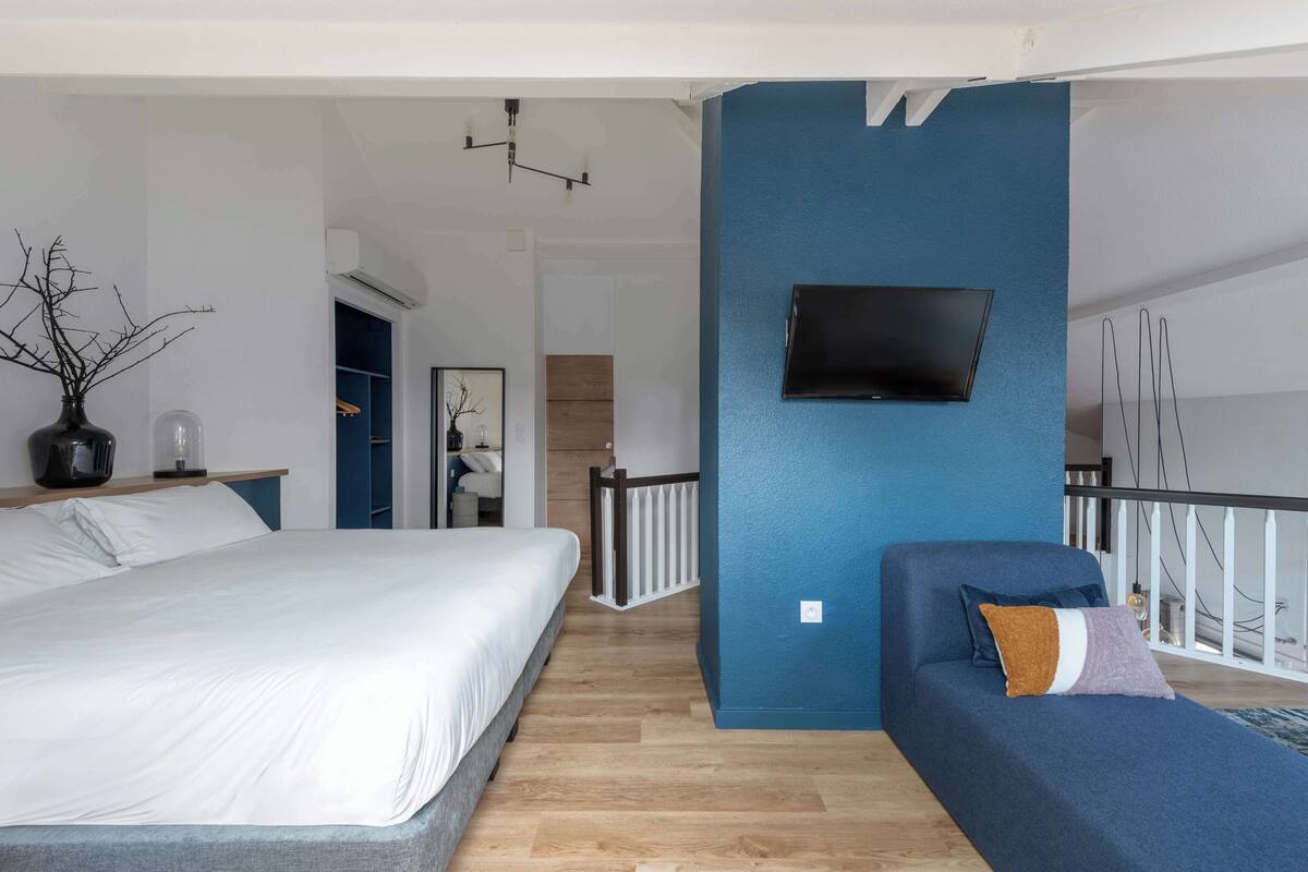 A modern bedroom with a large bed, a sofa, a TV mounted on a blue wall, and a balcony with a railing.