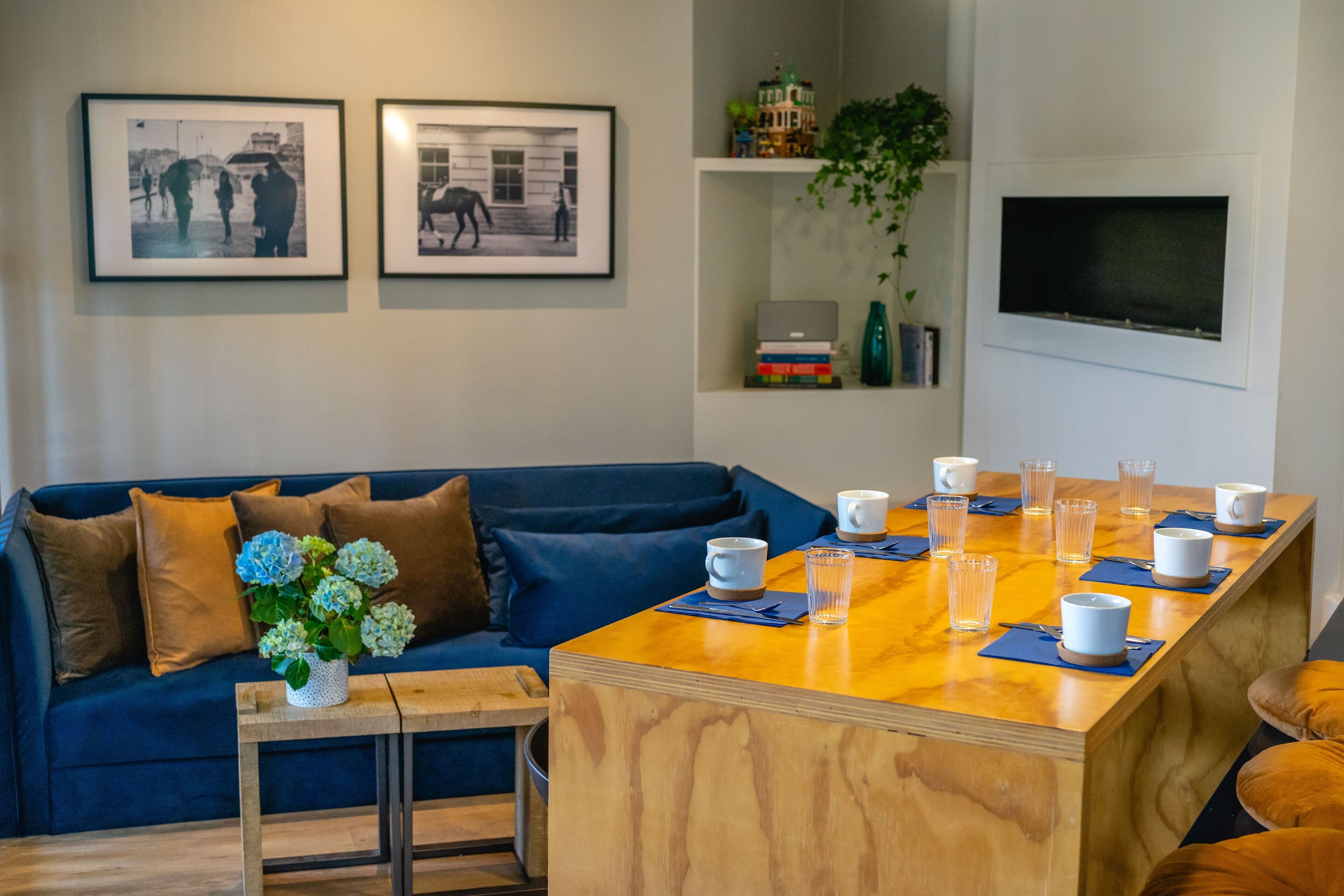 Een moderne woonkamer met een blauwe bank, een houten bijzettafeltje met bekers en glazen, zwart-wit foto's in lijsten aan de muur, en een tv boven het tafeltje.