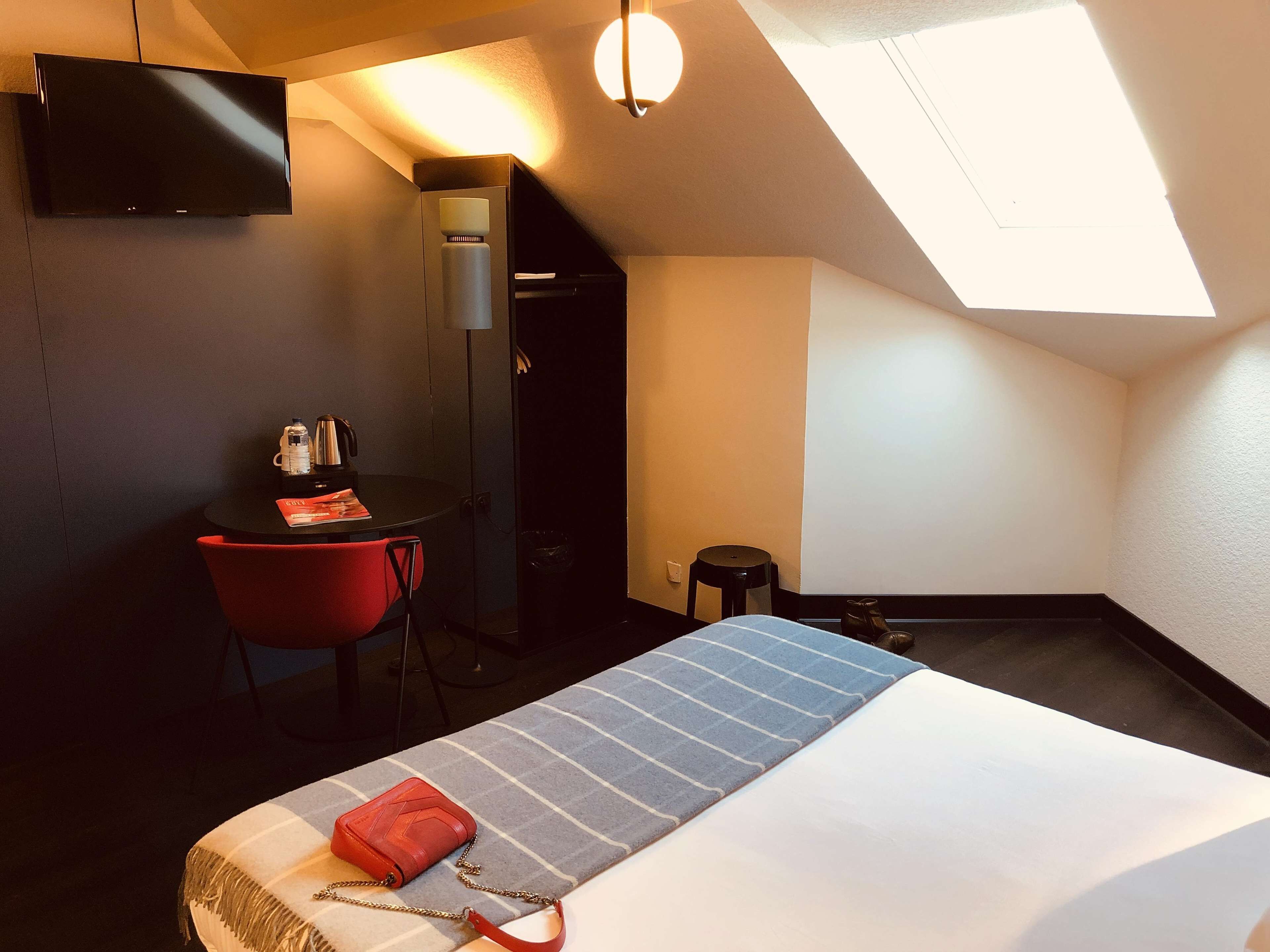 A hotel room with a bed, a desk, a chair, a small table, a TV, and a skylight.
