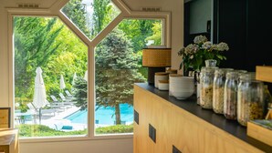 A view through a large window to a pool area with umbrellas and lounge chairs, a wooden counter with jars, and a lamp. - Best Western Bridge Hotel Lyon East (Jons)