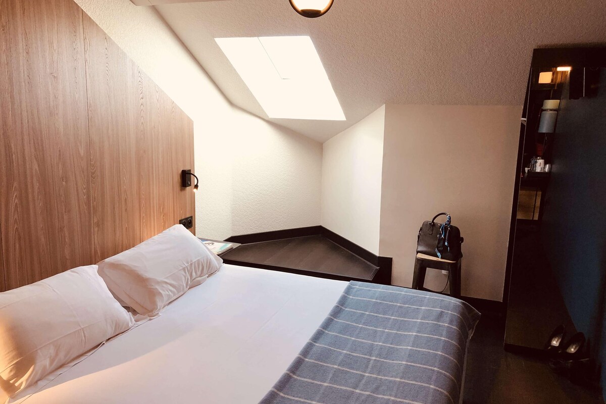 A bedroom with a bed, a skylight, a wooden wall, a small table, and a bag.